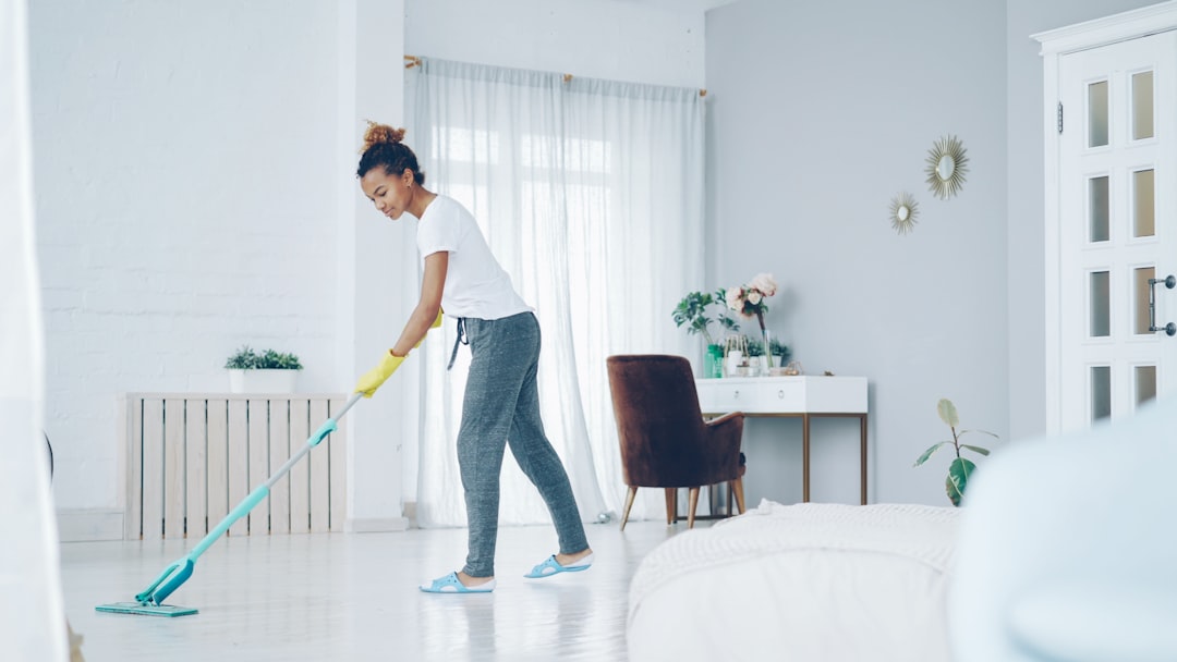 A pretty African American girl is mopping the floor at home with a flat plastic mop, wearing gloves and comfortable home clothing. Modern interiors, cleanliness, and young people concept.